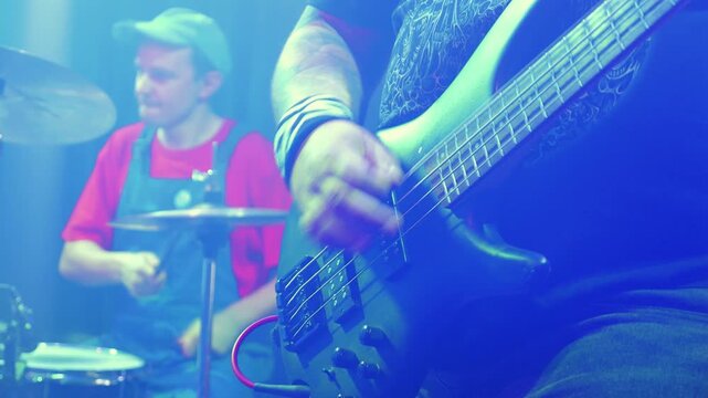 Close up of tattooed bassist playing bass guitar. Drummer in background at live concert. Energetic punk rock show with vibrant blue stage lighting and smoky atmosphere