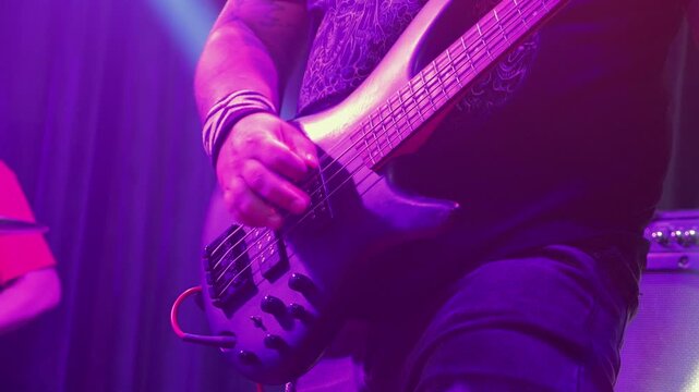 Close up of bassist playing electric guitar. Musician's hand has motion blur, conveying high energy. Stage is bathed in cool blue light with drummer in background. Raw, intense mood of live music