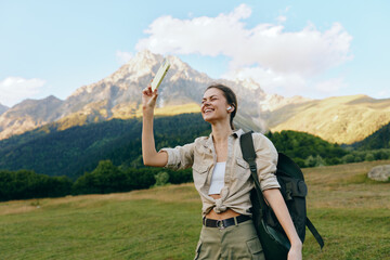 Fototapeta premium Young woman with a backpack stands in a grassy mountain meadow, holding a map up high and smiling as the sun shines on rugged peaks, signaling adventure
