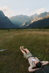 Fototapeta premium Woman relaxing in a grassy field near mountains, enjoying outdoor life, casual outfit, lying on her back with arms behind her head, serene natural scenery and peaceful mood