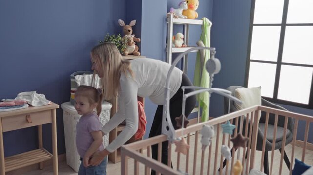 Woman handing a polka dot cloth to little girl reaching up at crib in nursery with shelves of toys and a wicker hamper visible; tenderness bonding.