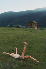 Fototapeta premium A young woman lies on a grassy field with one leg raised, expressing carefree energy as a bright mountain landscape and a distant building create a dramatic backdrop.