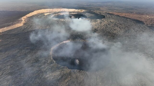 Aerial view of active volcanic craters with thick white smoke rising from the dark rocky terrain of the Danakil Desert Dalol, Afar, Ethiopia.