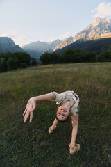 Fototapeta premium Young woman in a meadow outdoor scene performing a backbend, smiling at the camera, wind in hair, mountains in the background, colorful nature, relaxed lifestyle moment