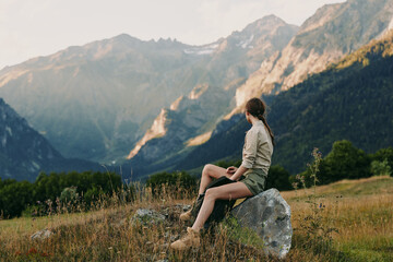 Fototapeta premium Woman sits on a rock in a mountain meadow, enjoying vast valleys and blue skies. Casual outdoor lifestyle shot emphasizing nature, wellness, and solitude.