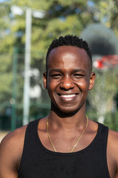 Vertical headshot portrait of happy young African American man in basketball court outdoors looking at camera smiling.
