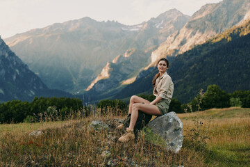 Fototapeta premium Young woman lifestyle sitting on rock in mountain meadow, casual outfit, relaxed pose, natural light, scenic landscape background, warm atmosphere