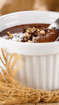 close up of creamy chocolate mousse in white ramekin topped with crushed hazelnuts, delicious homemade cocoa dessert with spoon