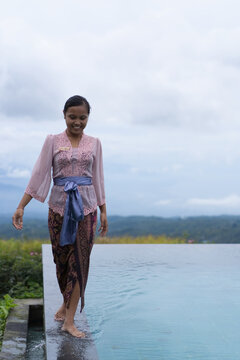 Smiling Balinese woman in kebaya walking along infinity pool, Bali