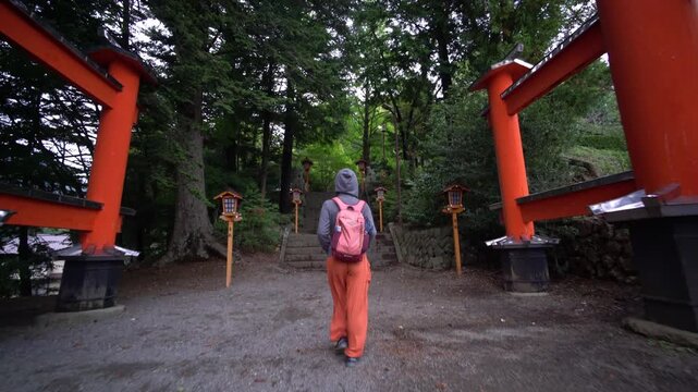 A female tourist with a backpack walks on a paved path towards the large red torii gate at the entrance of Arakura Fuji Sengen Shrine in Japan.