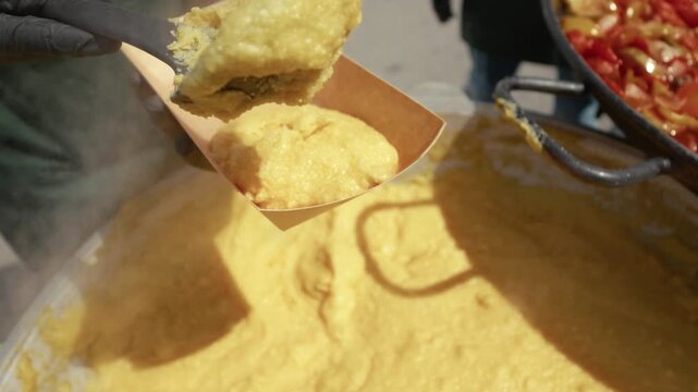 Chef scoops creamy yellow polenta with a wooden spatula from a large pot, while a pan of colorful vegetables simmers nearby in a vibrant outdoor kitchen setting