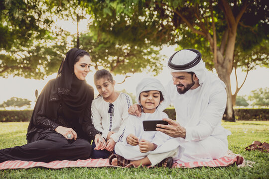 Arabian family in traditional dress smiling together on a picnic blanket in a sunny green park, parents and children enjoying leisure, bonding and relaxation outdoors