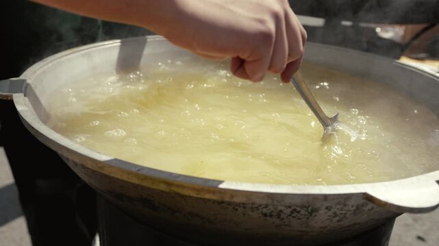 Person pours grain into a large pot of boiling liquid, then stirs the mixture with a spoon, showcasing the cooking process in an outdoor kitchen setting