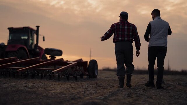 Farmer and agronomist walking in agricultural fields in sunset, back view. Agribusiness and agronomy, professionals discussing modern machinery for farming and soil processing in ecological region