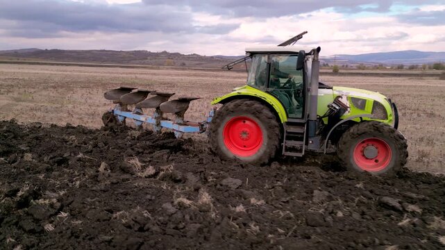 Aerial drone footage following a modern tractor pulling a mounted plow while turning soil across a large agricultural field. The clip highlights seasonal land preparation, mechanized farming, soil cul