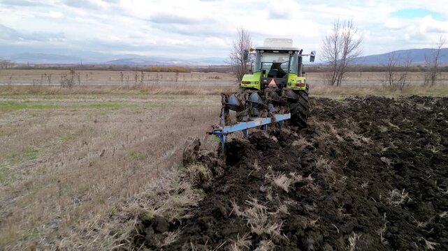 Aerial drone footage following a modern tractor pulling a mounted plow while turning soil across a large agricultural field. The clip highlights seasonal land preparation, mechanized farming, soil cul