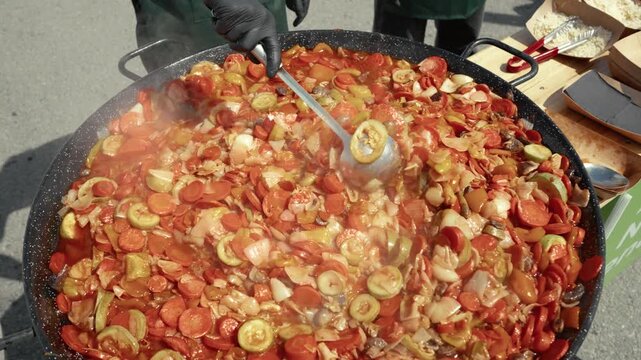 Chef in black gloves stirs a large pan of colorful vegetable stew with a ladle, steam rising, showcasing vibrant ingredients in an outdoor cooking setting