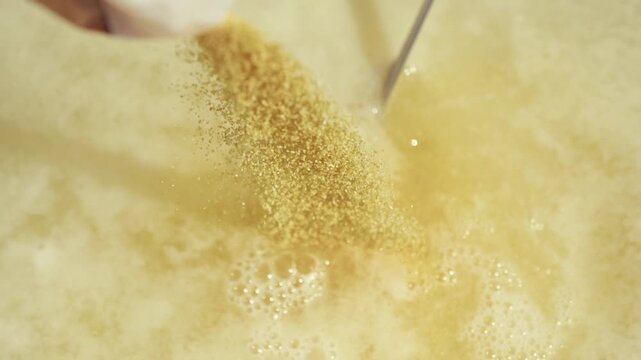 Hand pouring granulated sugar into bubbling liquid mixture, creating a frothy texture, with close-up focus on the sugar and foam in a clear container