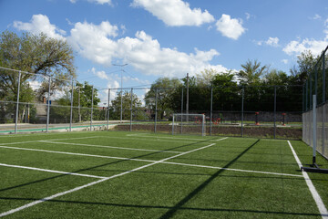 A wide shot of a community sports ground featuring green artificial grass, white markings, and a soccer goal under a blue sky with clouds. © Iuliana