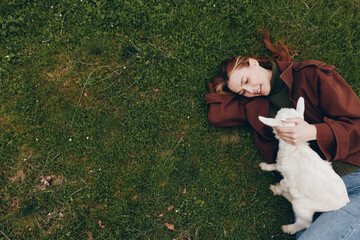 Fototapeta premium Cheerful outdoor moment of a woman lying on grass, cuddling a white goat kid, warm sunlight, relaxed posture, soft breeze, natural colors, candid lifestyle scene, gentle affection