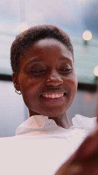 young Black woman laughing joyfully while on phone call. She is standing outdoors against a backdrop of string lights, creating a lively and vibrant atmosphere. Happy female enjoying call on cellular
