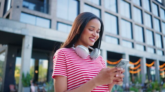 Smiling woman using smartphone with wireless headphones around neck. Female browsing mobile application while standing near modern apartments outdoors. Enjoying online communication during sunny day.