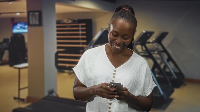 Young african american woman smiling and tapping smartphone while standing in a gym building; joyful connection.