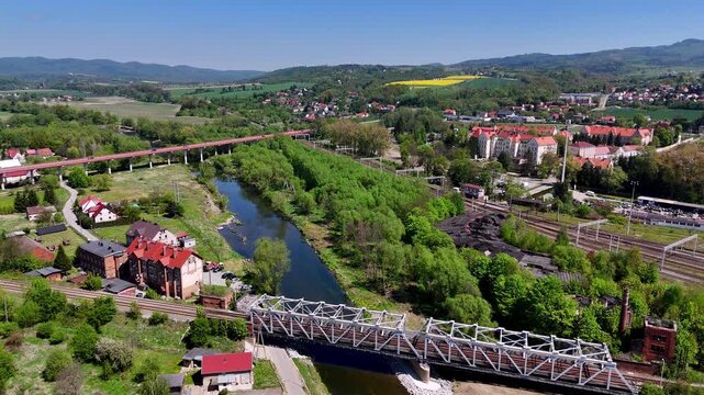 Aerial view of Kłodzko city, Nysa Kłodzka river flowing under a white steel railway bridge, red viaduct in the distance, urban buildings, railway tracks, green hills and rapeseed fields, sunny day, dr