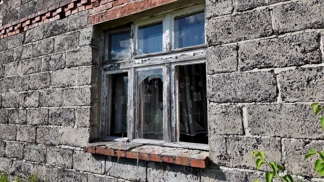 Old broken window with peeling white paint in a wall made of grey cinder blocks, shattered glass panes, dirty curtains visible inside, red brick windowsill, texture of damaged concrete and masonry, ru