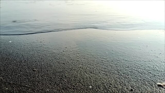 Gentle waves washing over dark sandy beach shoreline with calm water ripples, creating a peaceful tropical coastal atmosphere. Perfect natural background for relaxation and seaside themes