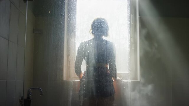 Female figure stands in bathroom, gazing out window through misted glass, sunlight streaming in, creating a serene atmosphere with soft shadows and reflections