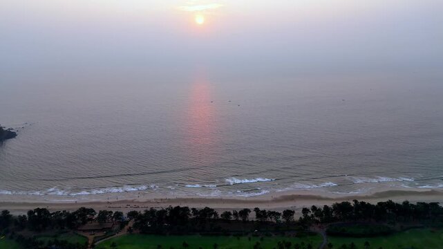 Drone shot capturing the tranquil coastline of Goa with fading sunlight glowing over the ocean, while rhythmic waves roll toward the sandy beach in a peaceful atmosphere.