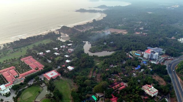 Cinematic aerial view of a tropical coastal landscape featuring luxury resorts, dense palm-covered greenery, winding water bodies, and a long sandy shoreline stretching beside the Arabian Sea.