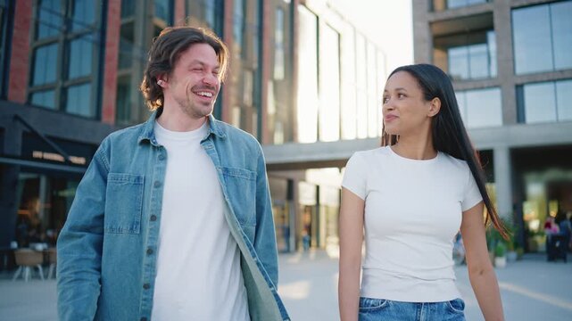 Smiling man and woman walking near modern downtown buildings outdoors. Friends talking while spending sunny afternoon together in busy city district. Urban relationship lifestyle concept.