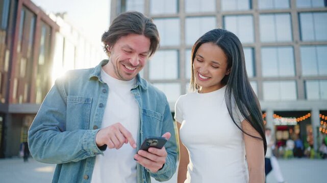Man showing smartphone screen to smiling woman near modern buildings. Friends checking mobile application together during sunny afternoon outdoors in busy downtown district. Technology concept.