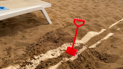 Red plastic shovel standing upright in sand near a white lounge chair on a beach, with footprints and a clear blue sky visible in the background © TatjanaMeininger