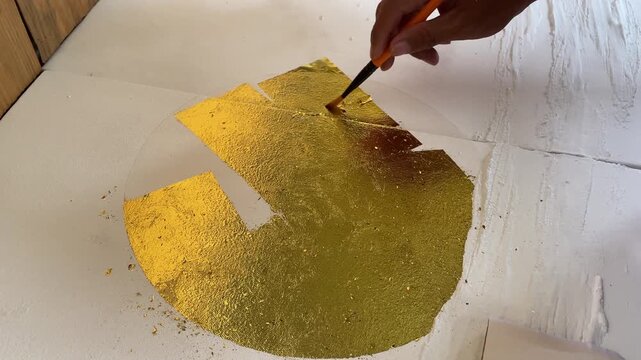 Handheld 4K close-up of hands applying shimmering gold leaf to a canvas. Selective focus capturing the artistic process of creating luxury textured wall art.