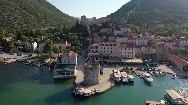A stunning drone shot of Mali Ston, featuring its historic stone fortress tower, red-roofed buildings, and peaceful harbor. The footage captures the charm of the Croatian coast and green mountains.