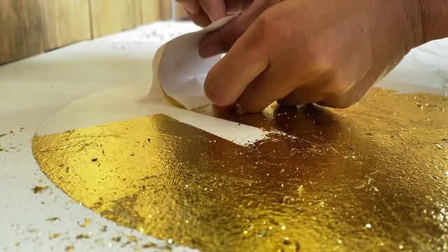 Handheld 4K close-up of hands applying shimmering gold leaf to a canvas. Selective focus capturing the artistic process of creating luxury textured wall art.
