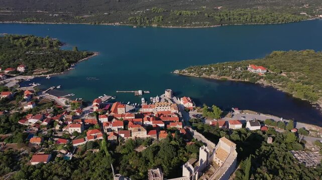 A stunning drone shot of Mali Ston, featuring its historic stone fortress tower, red-roofed buildings, and peaceful harbor. The footage captures the charm of the Croatian coast and green mountains.