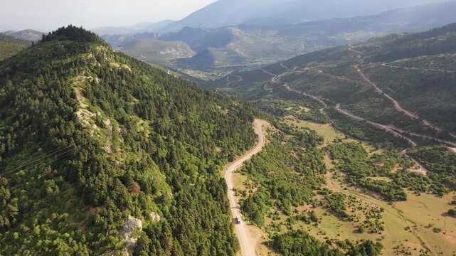 Stunning drone shot of a serpentine road cutting through the green, rocky mountainside of Delphi. The footage showcases the vast, wild landscape of Mount Parnassus under a bright, cloudy sky.