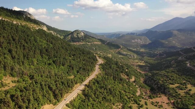 Stunning drone shot of a serpentine road cutting through the green, rocky mountainside of Delphi. The footage showcases the vast, wild landscape of Mount Parnassus under a bright, cloudy sky.
