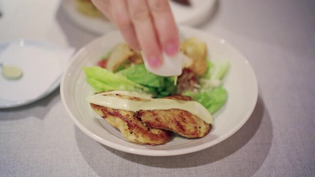 Close up of a woman squeezing creamy dressing from a small white sauce jug over grilled chicken breast served in a white bowl with Caesar style salad ingredients, on a light tablecloth