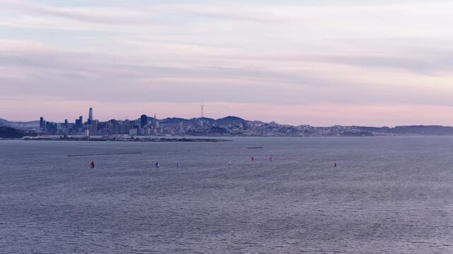 Stunning sunset view of the San Francisco skyline from across the bay, with sailboats dotting the water under a pastel-colored sky and the Sutro Tower visible in the distance