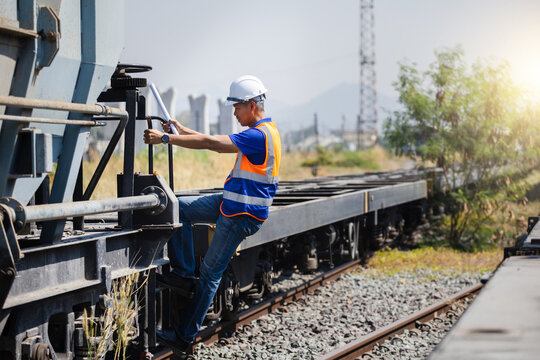 Railway engineer inspecting cargo train at industrial shipping yard, Worker man in safety vest climbing freight train for inspection, Logistics technician checking railway transport system on site
