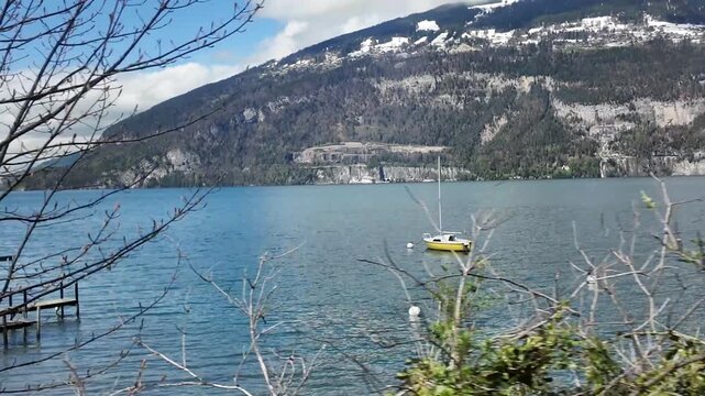 View from a moving train along Lake Thun from Interlaken to Zurich, featuring calm blue water, a sailboat, and alpine mountains in scenic Switzerland.
