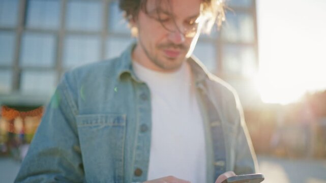 Closeup view of man using smartphone during sunny afternoon outdoors. Male browsing mobile application near modern downtown buildings at golden hour. Digital communication lifestyle concept.