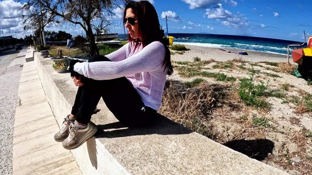 A woman sitting on the wall of a promenade in a small village overlooking the Mediterranean Sea. The woman has a sullen expression. The video was shot in the seaside village of Rodia, Sicily, Italy