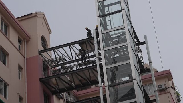 This photo shows construction workers installing an outdoor glass-paneled elevator and attached walkway on the exterior of a multi-story residential building.