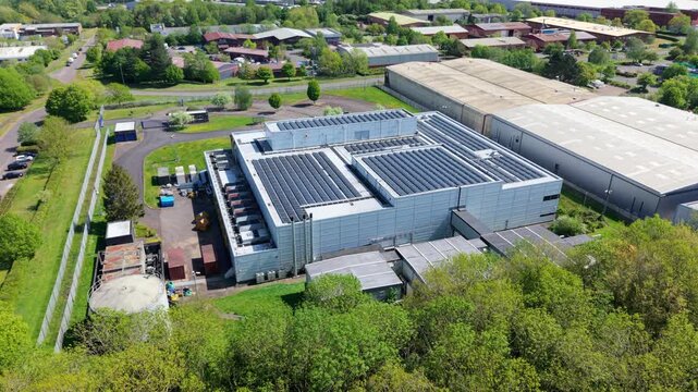 Aerial view of a large industrial data center with solar panel arrays on the roof surrounded by lush green trees and buildings in Wellingborough, England, United Kingdom.
